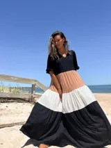 Vestido largo de diseño tricolor con bloques de color en negro, beige y crudo. Presenta escote en V, mangas cortas con volados y una falda amplia con cortes horizontales que crean un efecto escalonado.