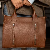 Cartera de cuero color tostado, estructurada, con doble asa de mano, correa desmontable y dos bolsillos frontales verticales con cierre. Presenta detalles de costuras pespunteadas y el logo de la marca grabado en la parte inferior.