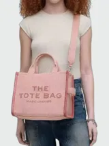 Bolso tipo tote de la marca Marc Jacobs, modelo The Medium Tote, confeccionado en lona color rosado. Presenta el texto "THE TOTE BAG MARC JACOBS" bordado en el frente, asas superiores y correa ajustable y desmontable para el hombro.