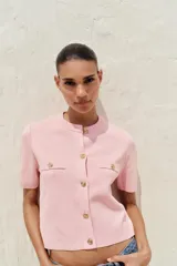 Chaqueta de punto color amarillo pálido, con cuello redondo, manga corta, cierre frontal con botones dorados y bolsillos de parche en el pecho.