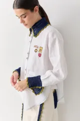 Camisa blanca de corte holgado con cuello y puños tejidos en crochet azul con borde verde. Presenta parches bordados decorativos en el frente y las mangas, cierre frontal con botones y un bolsillo de parche en el pecho.