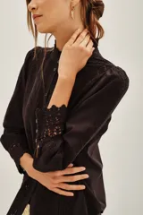 Camisa negra de algodón con cuello alto, cierre frontal de botones y detalles de encaje vertical y en los puños.