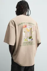 Camiseta de manga corta y cuello redondo, color beige claro, con corte relajado (relaxed fit) y un pequeño texto bordado en el pecho.