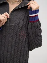 Pullover de algodón color beige, con tejido en ochos, cuello polo con cierre frontal y puños a rayas de colores (blanco, bordó y amarillo). El diseño es holgado y cae por debajo de la cadera.