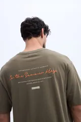 Remera oversize verde militar, confeccionada en 100% algodón, con cuello redondo y diseño relajado. Presenta un pequeño bordado frontal en tono naranja.