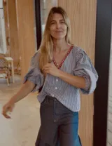 Camisa de rayas azules y blancas, de corte holgado con mangas abullonadas, escote en V y detalle de bordado rojo alrededor del cuello.