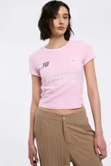 Remera baby tee de corte cropped y ajuste al cuerpo, color rosa con ribetes blancos en cuello y mangas. Presenta estampa frontal con el número 10, el año 94 y la frase 'GAME OVER' repetida tres veces.
