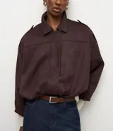 Campera bomber de gamuza sintética color marrón oscuro, con cuello clásico, cierre frontal, hombros caídos y puños ajustados. Presenta un diseño holgado con textura aterciopelada.