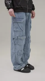 Pantalón tipo cargo de jean en color azul medio, con corte baggy fit y tiro bajo. Presenta múltiples bolsillos laterales con solapa y cierre de botón, además de un diseño holgado con frunces en la parte inferior de las piernas.