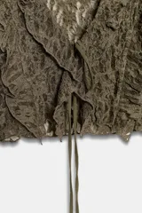 Chaqueta corta de tejido tipo encaje color verde oliva, con escote pico, manga larga acampanada y detalle de volantes en el borde del escote y los puños. Cierre frontal mediante lazadas.