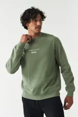 Buzo de felpa color verde oliva, con cuello redondo, puños y ruedo acanalados. Presenta un estampado frontal con texto en inglés y japonés junto al logo de la marca Mistral, y un detalle de etiqueta en la manga.