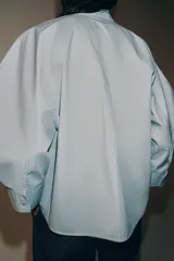 Camisa de algodón con cuello alto y mangas abullonadas por debajo del codo. Presenta cierre frontal con botones ocultos bajo solapa.