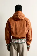 Cazadora de corte relajado para hombre, confeccionada en algodón con acabado efecto encerado en color marrón. Cuenta con cuello subido, capucha ajustable con cordones, mangas largas con puños elásticos y cierre frontal con cremallera. Incluye bolsillos frontales de vivo y forro interior a cuadros.
