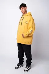Buzo canguro amarillo ocre oversize de algodón rústico con capucha, cordones negros con letras blancas, bolsillo frontal y puños y cintura acanalados. Presenta estampados en el frente y las mangas.