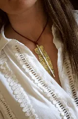 Collar con dije de bronce en forma de pluma alargada con textura perforada, suspendido de un cordón fino de color marrón.