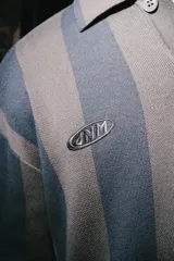 Chomba de manga larga con diseño de rayas verticales en tonos marrón y azul. Presenta cuello polo con cierre de botones y parches decorativos en el pecho.