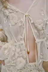 Blusa de escote en pico y manga larga con puños elásticos. Presenta un diseño semitransparente con bordados florales a tono, cintura elástica y cierre frontal mediante lazos con abalorios decorativos.