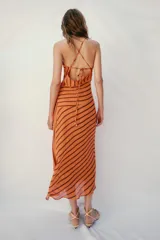Vestido midi de color naranja con rayas diagonales marrones, escote fluido y tirantes finos cruzados en la espalda con abalorios. Presenta corte evasé con abertura frontal y forro interior.