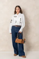 Camisa blanca de algodón, de corte corto y silueta holgada. Presenta cuello clásico, cierre frontal con botones y mangas largas con puños anchos. Está decorada con diversos parches y pines metálicos de estilo militar y náutico distribuidos en el frente y la manga derecha.