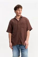 Camisa de manga corta color beige confeccionada en muselina de algodón, con cuello abierto, cierre frontal de botones y un bolsillo aplicado en el pecho.