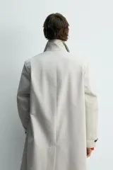Trench coat de corte relajado, color gris claro, confeccionado en tejido técnico con mezcla de algodón. Presenta cuello solapa, manga larga con puño ajustable, bolsillos de doble vivo en cadera, bolsillo interior y abertura en el centro de la espalda. Cierre frontal con botonadura oculta.