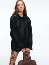 Chaqueta camisera de corte oversize en color negro, confeccionada en cuero sintético. Presenta cuello clásico, cierre frontal con botones y mangas largas con puños abotonados.