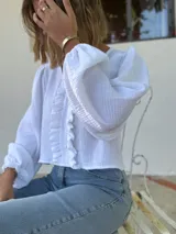 Blusa blanca de algodón con textura arrugada, cuello redondo, mangas largas abullonadas con puños elásticos y cierre frontal con botones y volantes.