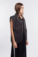 Chaleco de denim negro con efecto deslavado, diseño sin mangas, bolsillos frontales con solapa, costuras en contraste y botones metálicos.