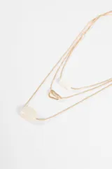 Collar triple de cadena fina plateada, con tres niveles de largo. El primer nivel presenta un dije cilíndrico blanco, el segundo un dije de mosquetón metálico y el tercero un dije de piedra blanca ovalada.