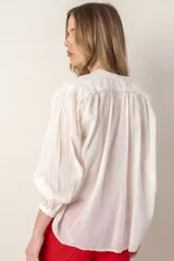 Camisa color crema de corte holgado, con cuello mao, cierre frontal con botones y volantes, mangas largas con puños elásticos y bordados en hombros y puños.