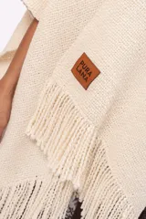 Ruana de tejido de punto en color crudo, con diseño abierto, flecos en el borde inferior y etiqueta de cuero con la marca Pura Lana en el frente.