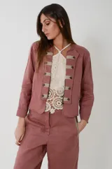 Chaqueta de lino estilo napoleónica color rosa palo, de cuello mao, manga larga y corte a la cintura. Cuenta con cierre frontal parcial de metal y detalle en la pechera con aplicación de cintas y botones decorativos.