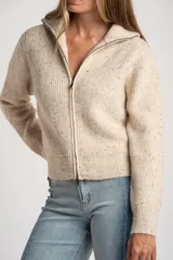 Cardigan de punto acanalado en color beige melange con motas oscuras. Presenta un diseño ajustado al cuerpo, cierre frontal completo con cremallera metálica y cuello alto.
