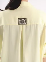 Camisa de manga corta color amarillo claro, con calce oversized, hombros caídos y cierre frontal con botones. Cuenta con bolsillos delanteros con bordados geométricos y mangas con dobladillo.