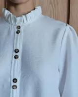 Camisa de manga tres cuartos color beige con cuello con volados y botones frontales. Presenta un detalle de color negro en la parte superior de las mangas.