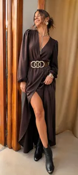 Vestido largo de satén color marrón, con escote cruzado en V, mangas largas abullonadas con puños elásticos y falda con tajo frontal. Incluye un cinturón ancho de cuero con hebilla metálica de tres aros entrelazados.