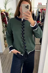 Saco tejido de punto color verde con cierre frontal de alamares y botones. Presenta cuello redondo, mangas largas con puños acanalados en contraste azul marino y blanco, y detalle de volados en el cuello y puños.