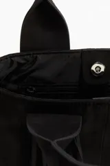 Bolso tipo shopper de color negro, confeccionado en tejido cepillado (similar a gamuza o nobuk). Presenta dos asas anchas con flecos largos colgantes en la parte frontal. Cierre magnético oculto en la parte superior y forro interior de tafeta con bolsillo con cierre.