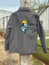 Campera impermeable de tela taslon color negro, con diseño de cuello alto y cierre frontal. Presenta un gran bordado en la espalda que incluye el escudo nacional uruguayo y el logo de la marca Bagual, además de un parche circular con la bandera de Uruguay en la manga.