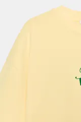 Camiseta amarilla de corte oversize, con cuello redondo, manga corta y estampa frontal en color verde con texto alusivo a Brasil Ipanema y pequeños símbolos.