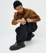 Pantalón cargo de corte mega baggy, color negro, confeccionado en sarga de algodón. Presenta múltiples bolsillos funcionales y un diseño versátil que permite transformar la prenda en bermuda mediante un sistema de ajuste en las piernas.