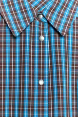 Vestido corto de manga larga con cuello solapa y cierre frontal de botones a presión. Presenta un diseño a cuadros en tonos azul y marrón, cintura ajustable con cinturón de hebilla y falda con volantes en el bajo.