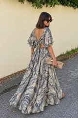 Vestido largo de diseño fluido y tacto sedoso, con estampado abstracto en tonos grises, beige y destellos dorados. Presenta escote en V en la espalda con detalle de lazo para atar, mangas cortas amplias con volados y falda larga con movimiento.