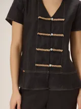 Camisa de manga corta color beige con estampado a cuadros, cuello en V y detalles de galones decorativos estilo oriental en la parte frontal.