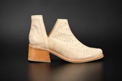 Botín de cuero con textura tipo crocco en color beige, con diseño de aberturas laterales en forma de V y detalles calados en el empeine. Cuenta con un taco cuadrado de madera foliada y suela de goma antideslizante.