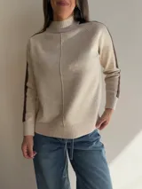 Sweater de tejido de punto en color beige con cuello alto y costura central vertical. Presenta mangas ranglan con una franja lateral en color marrón oscuro que genera contraste, y terminaciones en tejido acanalado en puños y ruedo.