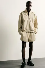 Chaqueta estilo deportivo de corte relajado, color beige con detalles de vivos en color verde oscuro. Presenta cuello alto, cierre frontal con cremallera de doble dirección, bolsillos laterales y puños elásticos.