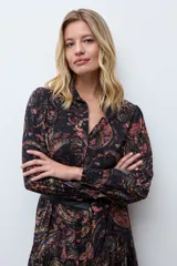 Vestido midi de corte camisero con estampado paisley en tonos oscuros, manga larga con puños ajustados y cierre frontal de botones. Presenta un diseño fluido con falda de volantes en el ruedo.