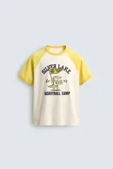 Camiseta de corte relajado con mangas raglán cortas en color amarillo y cuerpo en color crema. Presenta un estampado gráfico frontal con temática de campamento de básquetbol y un cocodrilo.