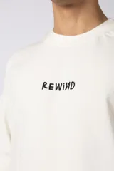 Remera negra de corte oversize confeccionada en tela pesada. Presenta un bordado frontal con la palabra 'REWIND' y un diseño de parche en la espalda con estética de papel roto.